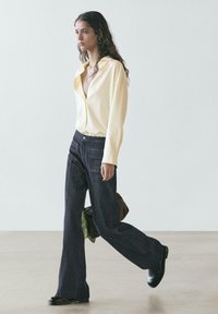 Langärmliges, hellgelbes Hemd mit Knopfleiste, kombiniert mit dunklen, ausgestellten Jeanshosen. Merkmale sind eine lockere Passform und Seitentaschen.