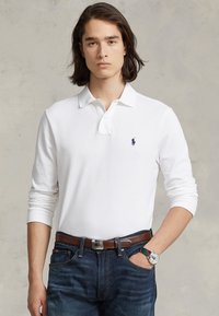 Witte lang/lang mouw polo met een kraag en een klein marineblauw logo op de linkerkant van de borst, gecombineerd met donkere jeans en een bruine riem.