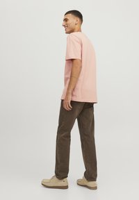 Lichtroze katoenen T-shirt met een ontspannen pasvorm, gecombineerd met een bruine broek. De persoon staat met een neutrale achtergrond, wat casual kleding toont.
