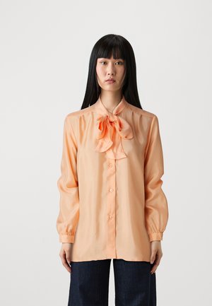 Peach-colored blouse in satin fabric, featuring a bow tie at the neckline, button front, and long, billowy sleeves with cuff detailing.