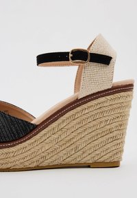 Sandalia de cuña con suela de yute trenzada, tira de tela negra, parte superior texturizada beige y detalle de hebilla dorada en el tobillo.