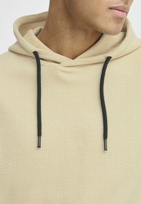 Beige hoodie i vaffelstruktur med snøre, der har sorte spidser. Nærbillede, der fremhæver materialet og detaljerne ved halsudskæringen.