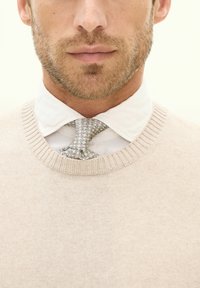 Beiger Pullover mit geripptem Ausschnitt, getragen über einem weißen Hemd mit Kragen. Eine gemusterte graue Krawatte ist am Kragen gebunden und verstärkt den Lagenlook.
