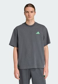 Camiseta gris de manga corta con cuello redondo, que presenta un logotipo verde de tres rayas en el pecho y una pequeña etiqueta verde en el dobladillo.