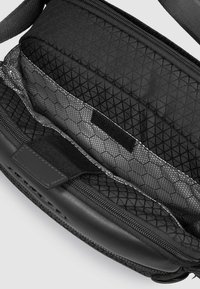 Interior de bolsa negra con cremallera, con patrón geométrico texturizado, bolsillos de malla y una correa de velcro para asegurar los objetos. Base negra y lisa.