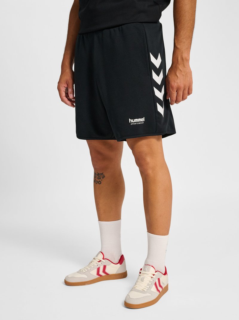 Mann trägt schwarze Hummel-Sportshorts mit weißen Chevron-Streifen, kombiniert mit weißen Socken und beige-roten Sneakers.