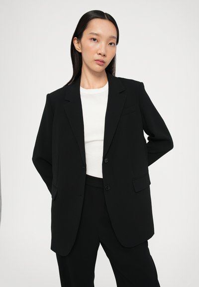 Schwarzer maßgeschneiderter Blazer mit zwei Knöpfen, Revers und seitlichen Taschen. Getragen über einem weißen Hemd, mit einer glatten, strukturierten Silhouette.