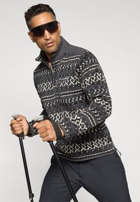 Pull-over en polaire à motif noir et blanc avec col zippé et logo. Pantalons sombres et bâtons de trekking tenus dans les mains, avec des lunettes de soleil.