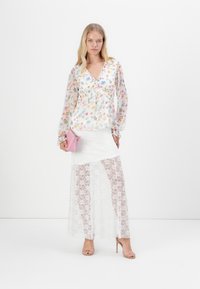 Blusa transparente con estampado floral y mangas largas, combinada con una falda blanca de encaje con una sobrecapa transparente. Un pequeño bolso rosa realza el conjunto.