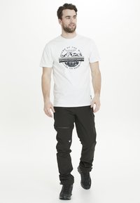 Vit grafisk t-shirt med bergdesign och svart text, tillsammans med svarta utomhusbyxor som har blixtlåsfickor och mönstrad textur.