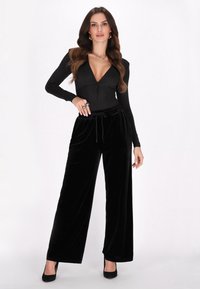 Fekete, hosszú ujjú body mély dekoltázzsal, kombinálva magas derekú fekete velúr, bőszárú nadrággal és fekete magassarkú cipővel.