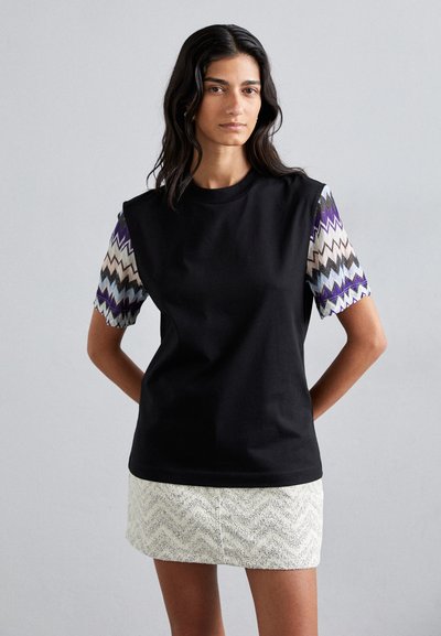 T-shirt en coton noir à manches courtes et motif présentant un design en zigzag violet, bleu et crème. Col rond simple. Porté avec une jupe blanche texturée.