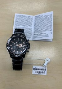Zwarte chronograaf horloge met een glanzende afwerking, roségouden accenten, multi-dial weergave en een label voor garantie-informatie naast een handleiding.