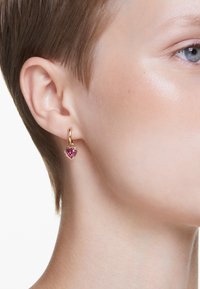Brincos de argola dourados com uma pedra preciosa em forma de coração rosa. Vista em close destacando sua forma e acabamento brilhante contra o tom de pele.