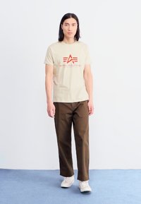 Beige cotton t-shirt with red logo and text "ALPHA INDUSTRIES," paired with dark brown trousers and white sneakers. Plain background.