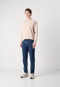 Beige pulóver kerek nyakkal és finom folt részlettel, kék farmernadrággal és világosszürke sportcipővel párosítva, semleges háttér előtt.