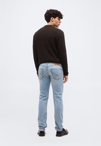 Άνδρας που φοράει σκούρο καφέ πουλόβερ, ανοιχτό μπλε τζιν και μαύρα παπούτσια, στέκεται στραμμένος ελαφρώς προς τα δεξιά πάνω σε απλό φόντο.