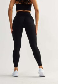 Schwarze hochgeschnittene Leggings mit glatter Textur und eng anliegendem Design, kombiniert mit weißen Sportschuhen. Keine sichtbaren Muster oder Logos.