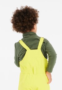 Lys gule overalls med justerbare stropper, båret over en mørkegrøn fleece-top. Bagviewet viser tekstur og elastisk talje-detail.