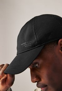 Cappellino nero con pannello frontale solido e retro in rete, cuciture visibili e logo del marchio bianco sul lato.