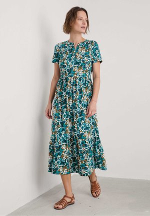 Rochie florală cu mâneci scurte, fundal teal, flori albe și maronii, talie strânsă și fustă cu volane. Asortată cu sandale maroniu cu barete.