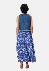 Denim cropped vest met rafelige zoom over een blauwe bloemenmaxirok met witte tropische patronen, gedragen met bruine instapschoenen.