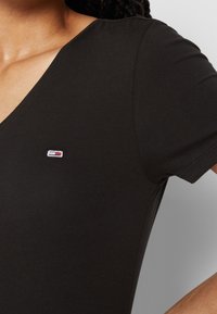 Närbild av en persons axel som bär en svart t-shirt med v-ringning som har en liten logo på bröstet, vilket framhäver tygstrukturen.