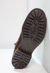 Brun lädersko med en texturerad gummisula som visar djupa räfflor och korsmönster, visad från botten mot en ljus bakgrund.