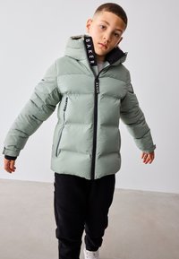 Jongen draagt een lichtgroene donsjas met capuchon, zwarte broek en witte sneakers, staat met licht gespreide armen.