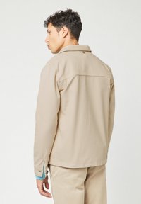 Beige lightweight jacket with a collar, button closure, and side pockets. Features contrasting blue accents at the cuffs and a smooth texture.