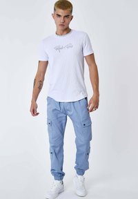 Giovane uomo con capelli biondi corti, che indossa una maglietta bianca "Project x Paris", pantaloni cargo di un blu chiaro e sneaker bianche, in piedi davanti a uno sfondo bianco.