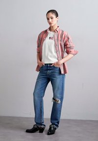 Camisa a cuadros en rosa y blanco, camiseta blanca, jeans desgastados azul, zuecos negros con acentos metálicos, de pie contra un fondo neutro.