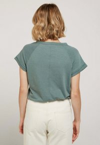 T-shirt à manches courtes, col ras du cou en teal atténué, fabriqué à partir d'un tissu doux avec des manches retroussées. Porté avec un pantalon taille haute de couleur claire.
