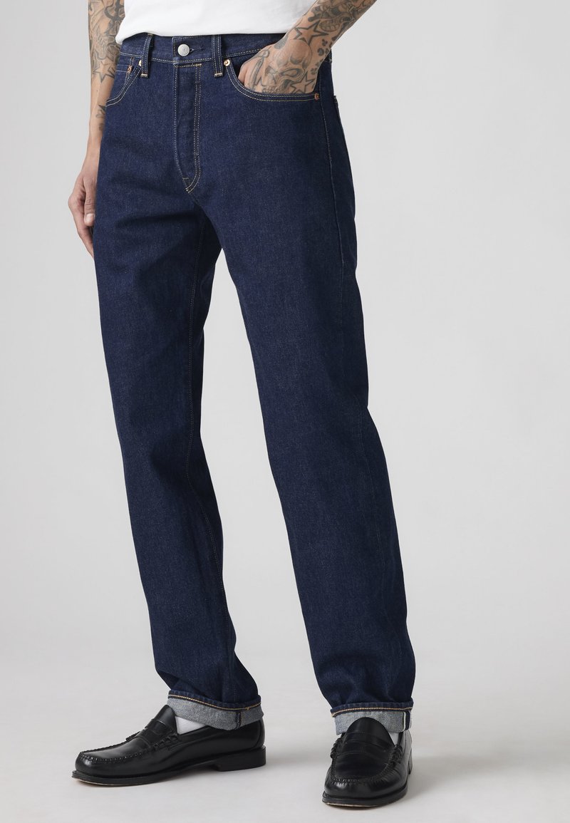Hombre con vaqueros azul oscuro de pierna recta con puños, calcetines blancos, mocasines negros y camisa blanca, manos en los bolsillos, brazos tatuados visibles.