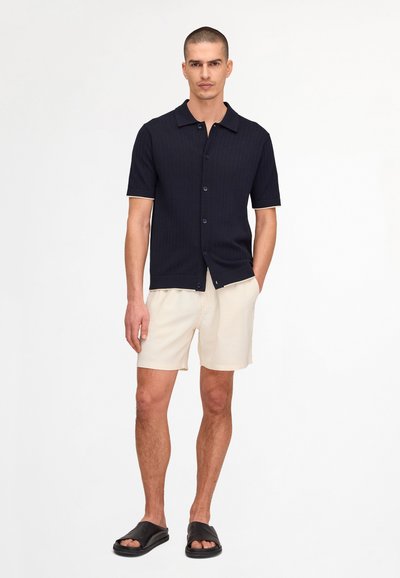 Man wearing a navy short-sleeve button-up shirt, beige shorts, and black slide sandals standing with one hand in pocket.