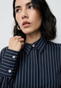 Marineblauw overhemd met witte verticale strepen, voorzien van een klassieke kraag en geruite manchetten. Glad stof met een frisse textuur.