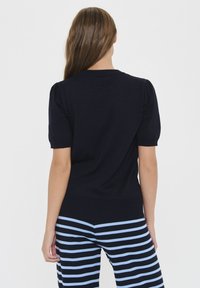 Jūras spēku tumši zils adīts tops ar īsām puffīgām piedurknēm un ribotu apakšu, savienots ar zilām un tumši zilām svītrainām biksēm. Gluda tekstūra, klasiskā dizaina.