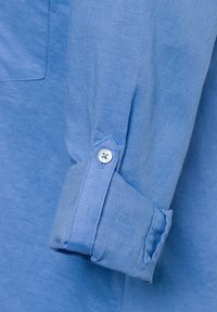 Camicia in lino azzurro chiaro con un solo bottone al polsino, caratterizzata da una texture morbida e un delicato motivo intrecciato vicino alla tasca.