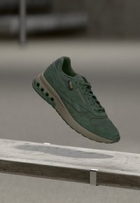 Grön atletisk sneaker med beige sula som har cirkulära luft hål, svävande över en betongbänk i en utomhusmiljö.