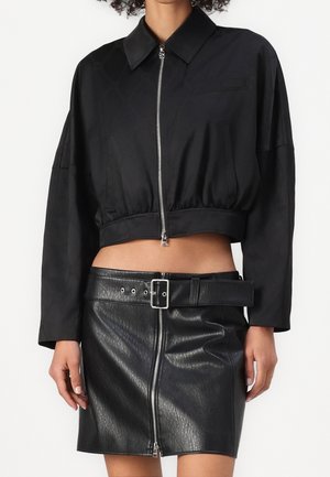 Mujer con chaqueta negra corta con cremallera y mini falda de cuero negra con cinturón ancho y cremallera frontal.
