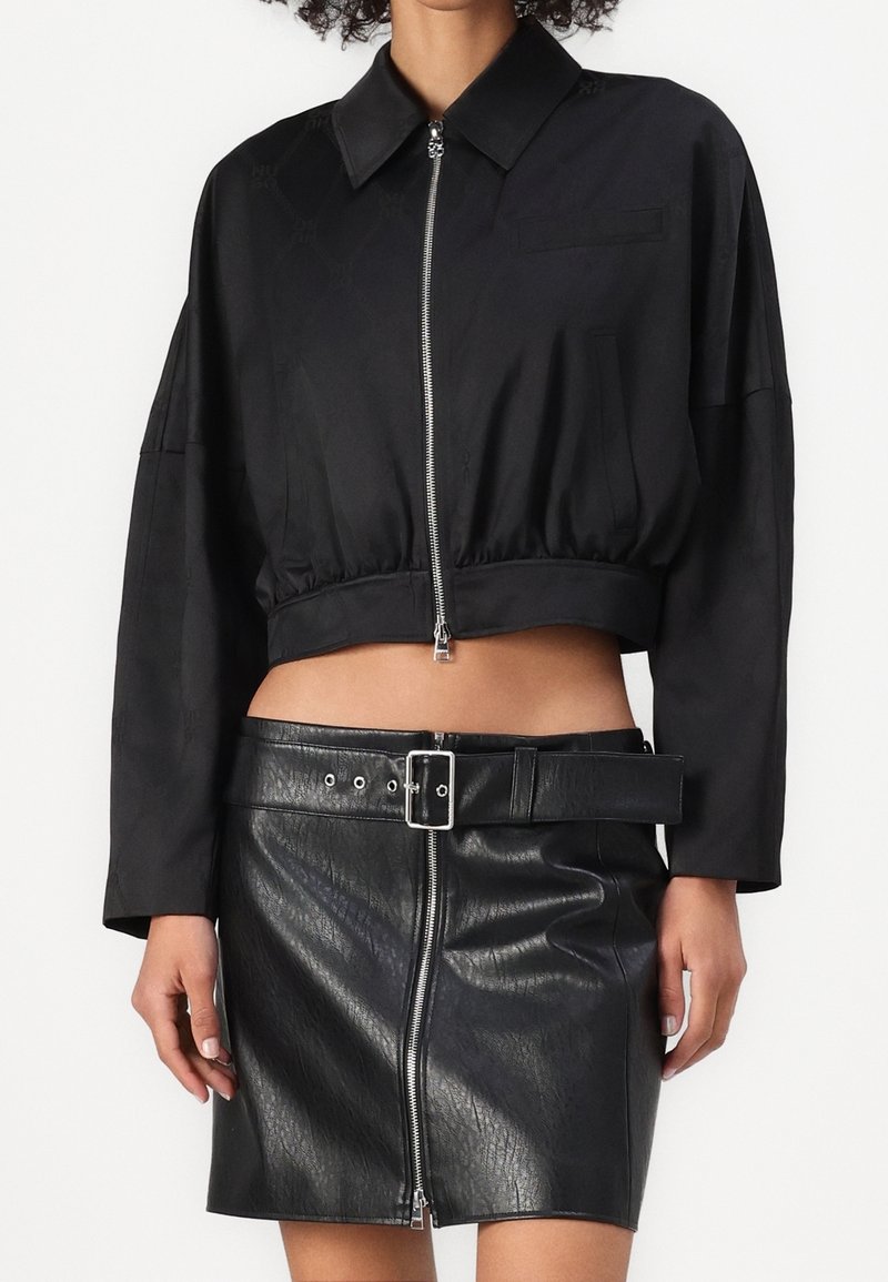 Femme portant une veste courte noire zippée et une mini-jupe en cuir noire avec une large ceinture et une fermeture éclair à l'avant.