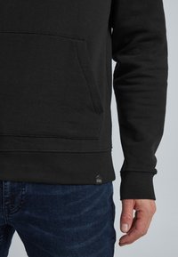 Man in zwarte hoodie met kangoeroezak en donkerblauwe spijkerbroek, hand ontspannen aan de zijkant tegen een neutrale achtergrond