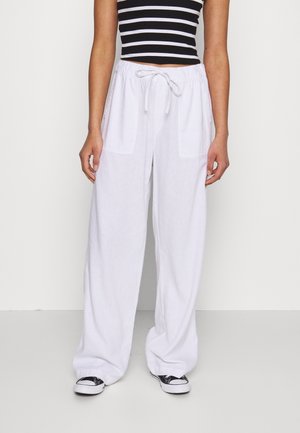 Person iført løstsiddende hvide bukser med snøre, sort-hvid stribet cropped top og sorte lavtop-sneakers, stående mod en ensartet baggrund.