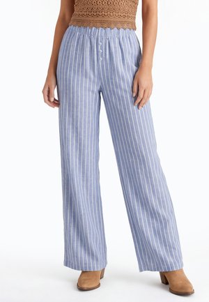 Vrouw draagt losse lichtblauwe broek met witte verticale strepen, een cropped bruin kanten top en beige laarzen met hak, staand tegen een witte achtergrond.