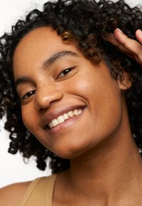 Personne souriante avec des cheveux bouclés mettant en avant leur texture naturelle. Peau lisse avec des taches de rousseur, portant un haut beige. L'arrière-plan est blanc uni.