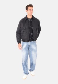 Giubbotto bomber nero, tessuto leggero, chiusura con zip, due tasche frontali, indossato sopra una camicia nera a fantasia e jeans larghi azzurri, sneakers bianche.