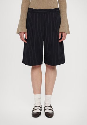 Laivastonsiniset pinstripe-shortsit, joissa on poimutukset, tasainen vyötärö ja sivutaskut, yhdistettynä valkoisiin sukkiin ja tummiin, kaksivöisiin kenkiin.