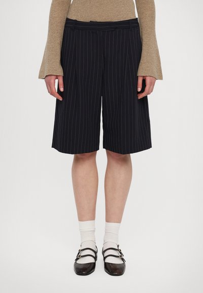Shorts bleu marine à rayures avec des plis, une taille plate et des poches latérales, associés à des chaussettes blanches et des chaussures sombres à double bride.