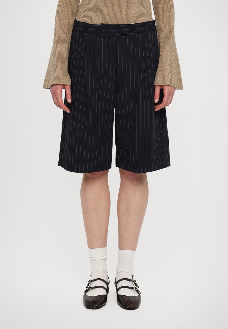 Marine-blå stripeshorts med stikninger, en flad talje, og sidelommer, parret med hvide sokker og mørke sko med dobbelt strop.