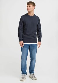 Marineblauer Pullover mit Rundhalsausschnitt, gerippten Manschetten und Saum, kombiniert mit hellblauen Jeans und grauen Sneakers mit beigen Akzenten. Schlichter, lässiger Look.